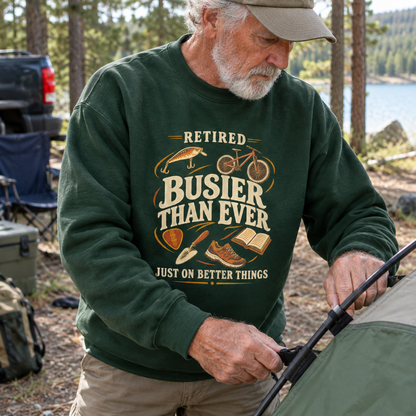 Senior man wearing dark green vintage sweatshirt with printed retirement slogan beside lakeside campsite.