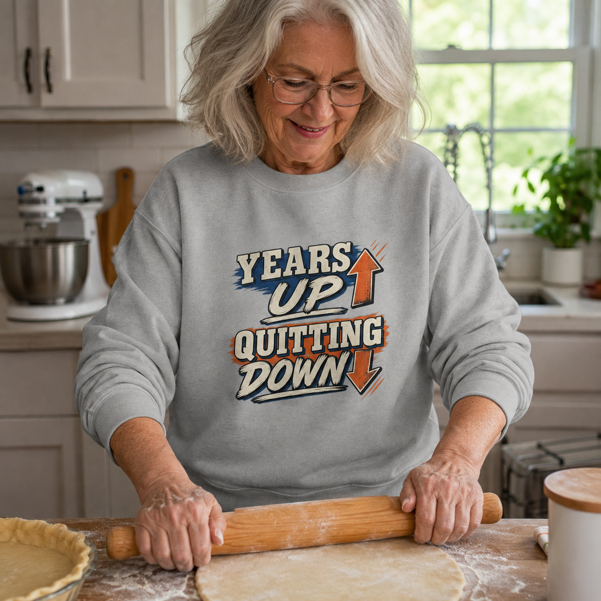 Gray vintage sweatshirt with printed “Years Up. Quitting Down” slogan, senior woman baking in kitchen.