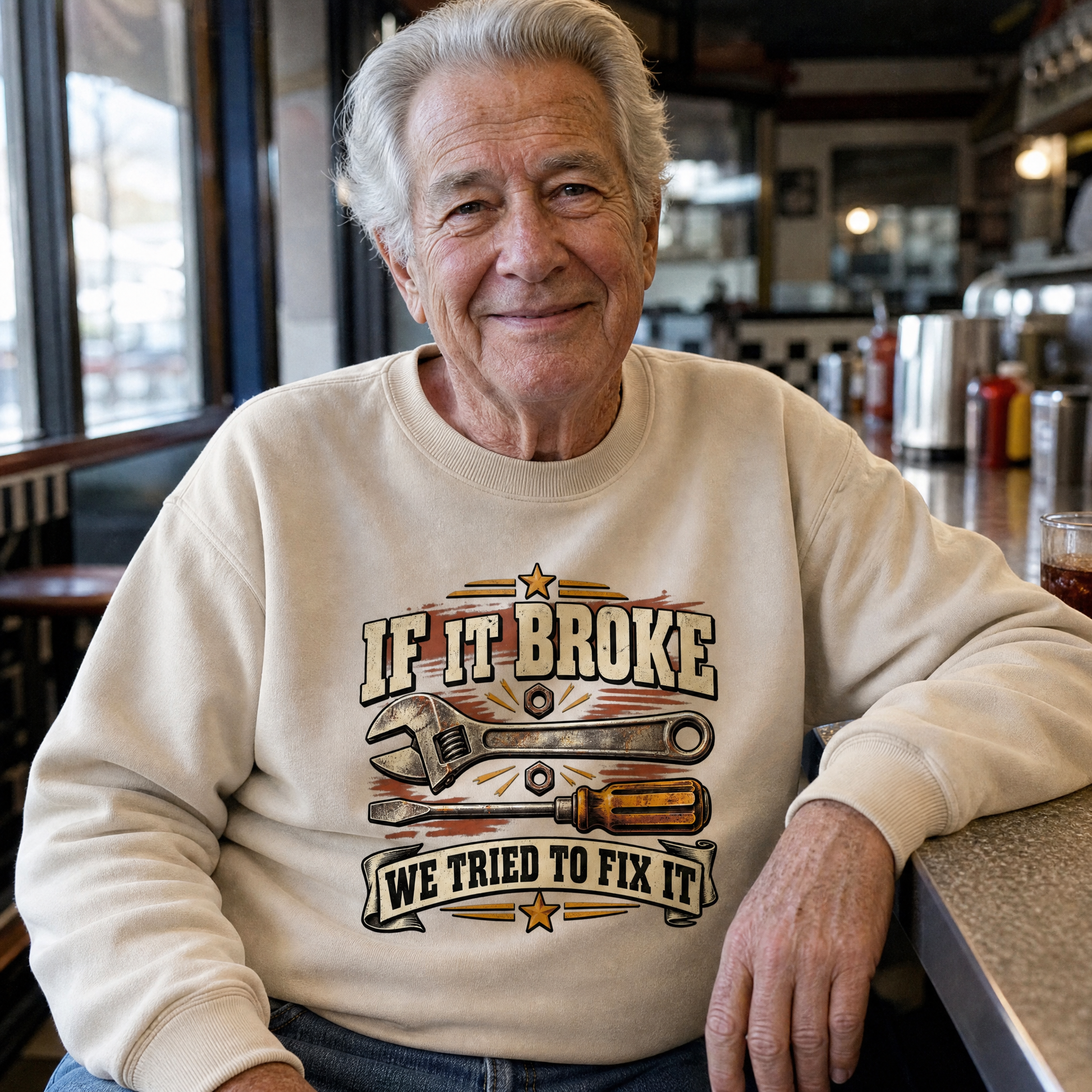 Senior man wearing cream vintage sweatshirt with printed “If It Broke We Tried To Fix It” design