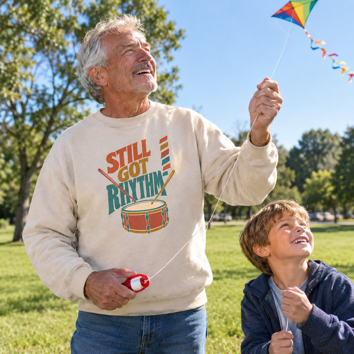 Senior man wearing cream vintage sweatshirt with printed Still Got Rhythm design, flying a kite outdoors.