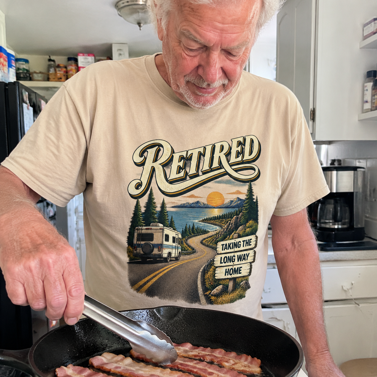 Beige vintage t-shirt with Retired Taking The Long Way Home print, senior man cooking indoors