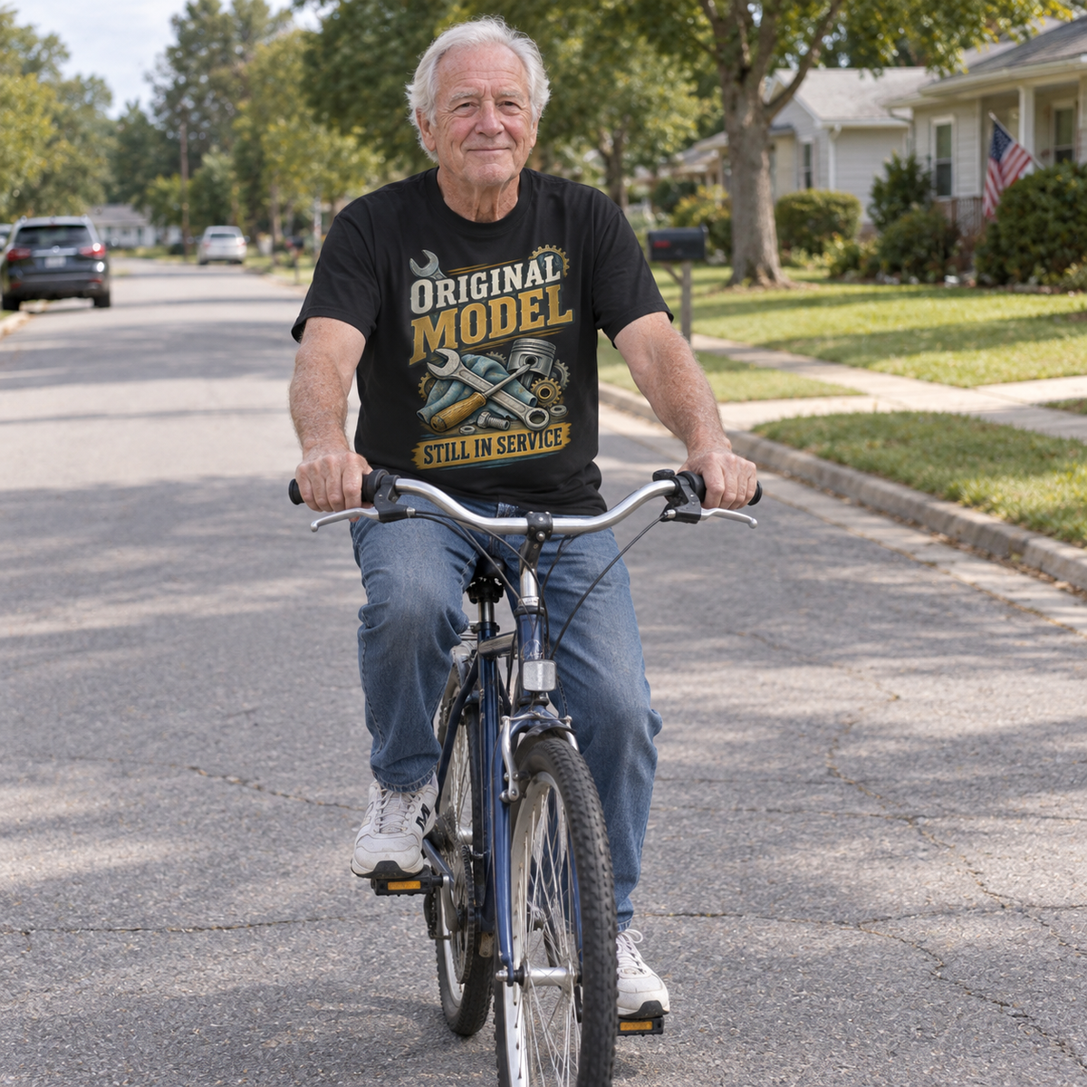 Senior man rides bicycle wearing black vintage t-shirt printed with Original Model Still In Service