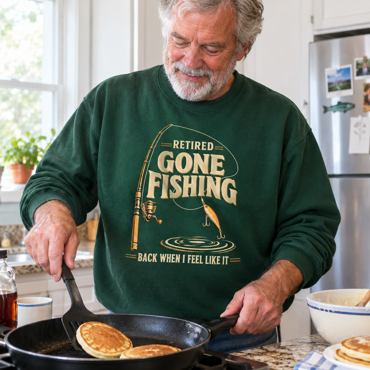 Dark green vintage sweatshirt with printed fishing slogan worn by senior man cooking pancakes in kitchen.