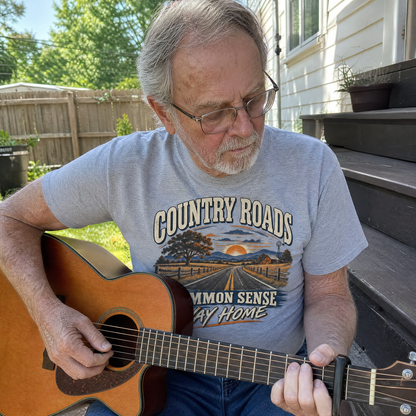 Senior man wearing gray vintage t-shirt with Country Roads slogan, playing guitar outdoors.