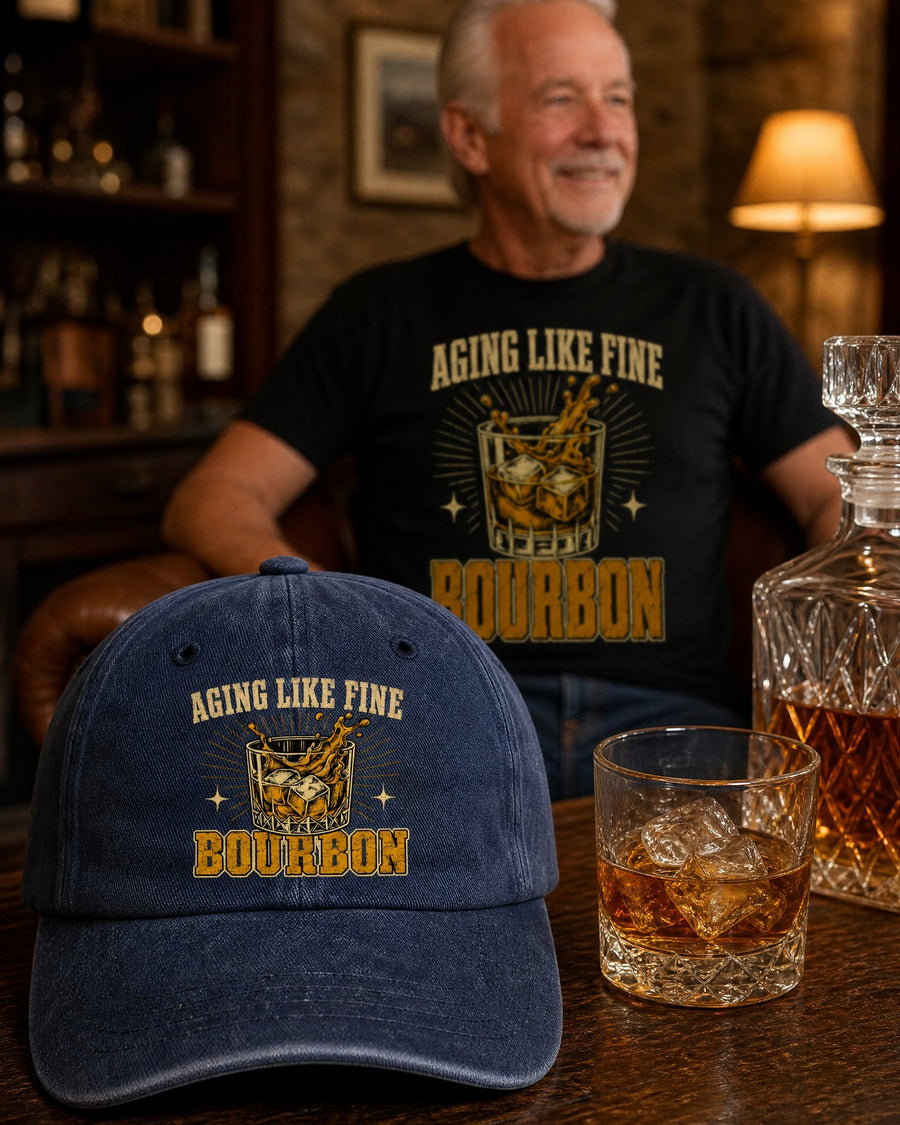 The navy Aging Like Fine Bourbon cap on a home bar beside a glass of bourbon, a man in the black tee behind