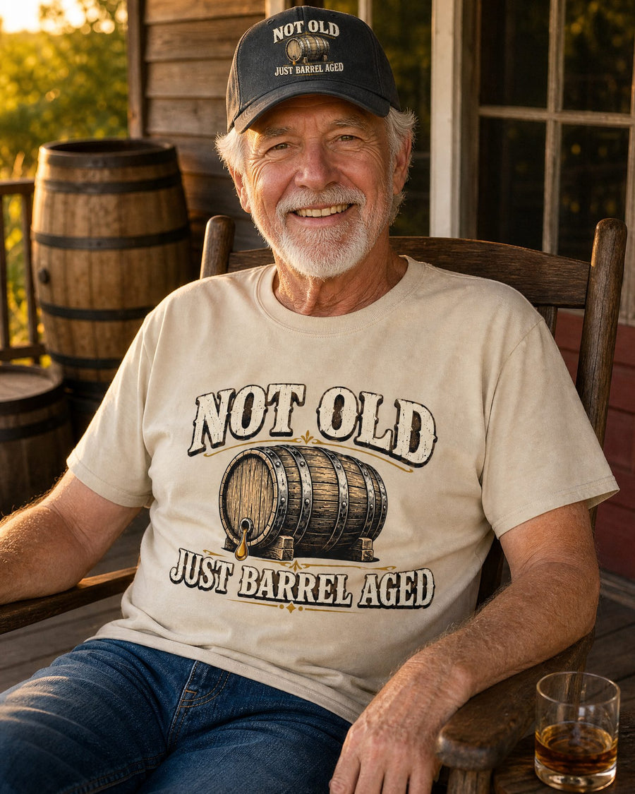 Man in the Not Old, Just Barrel Aged sand tee and black cap in a rocking chair on the porch