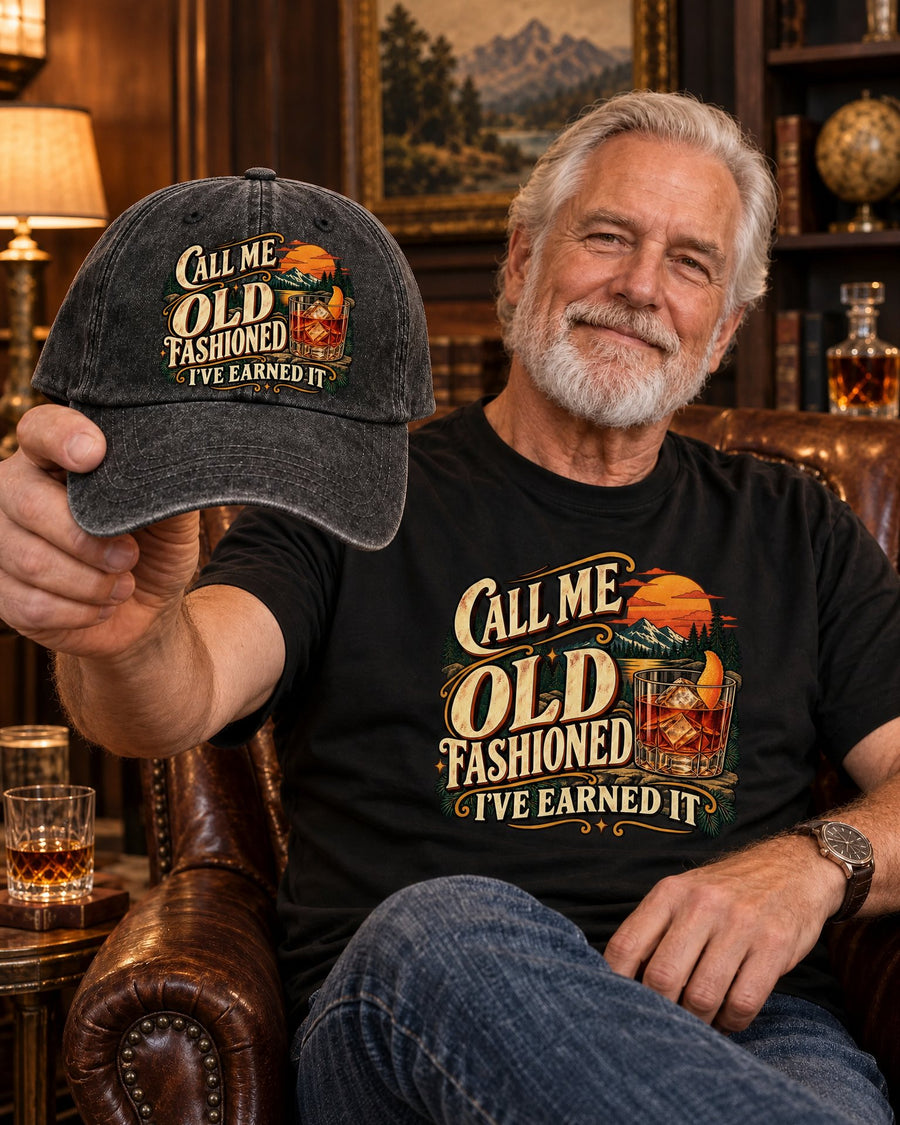 Man in a leather armchair in the black Call Me Old Fashioned tee, holding up the matching cap