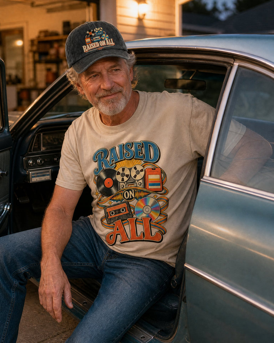 Man in the Raised On All sand tee and black cap sitting in his parked car with the door open