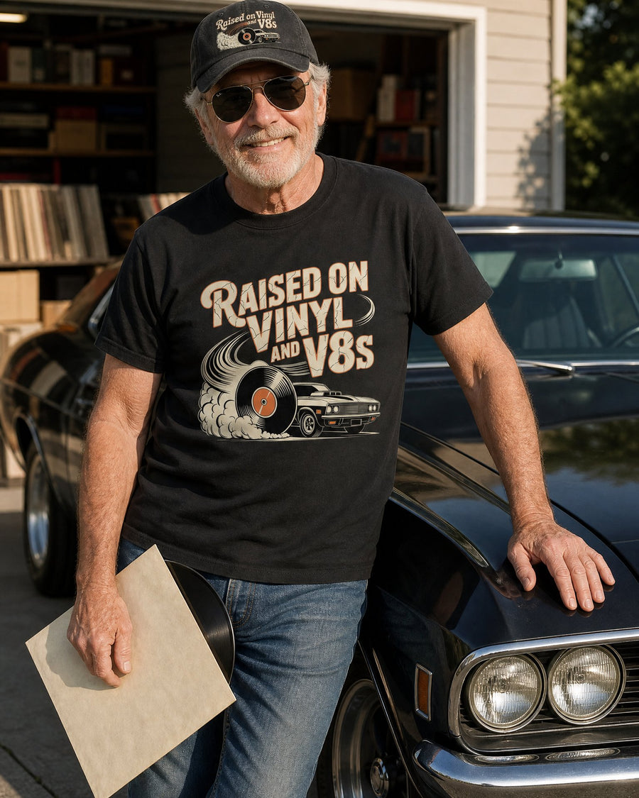 Man in the Raised on Vinyl and V8s black tee and black cap leaning on his classic car, holding a record